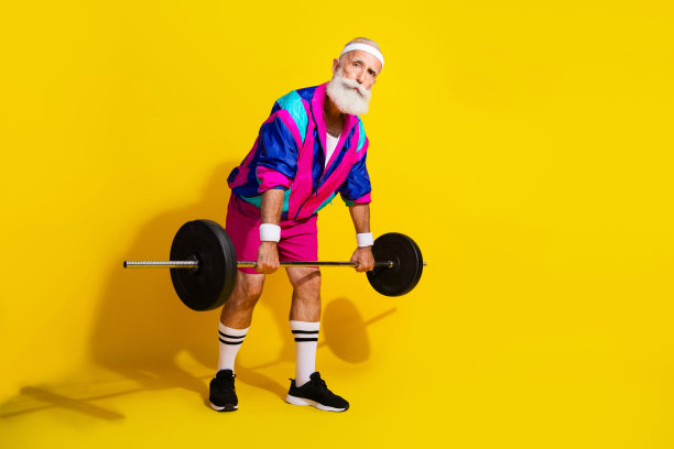 全身照片横幅，搞笑的老年运动员穿着时尚复古运动服，举起杠铃，背景为黄色。图片下载
