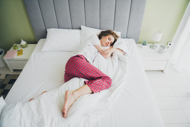 俯视照片中，穿着可爱夜衣的女孩微笑着，享受躺在室内公寓房间的床上。图片下载