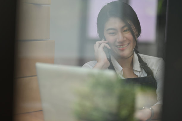 透过玻璃窗的照片，年轻女性中小企业主在店内与客户通话，同时工作，在线购物和配送概念。图片下载