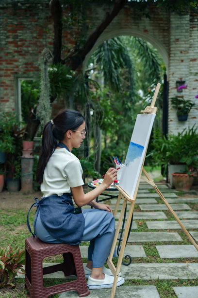 亚洲女性艺术家专注于在花园中绘制风景画。她使用水彩画，手握画笔，神情专注，创作出宁静的艺术作品。周围环绕着郁郁葱葱的绿意。图片下载
