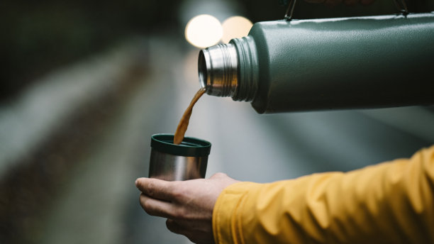 男人手部特写，从绿色保温瓶中倒咖啡到杯子里。金属不锈钢保温瓶在森林中的照片。图片下载