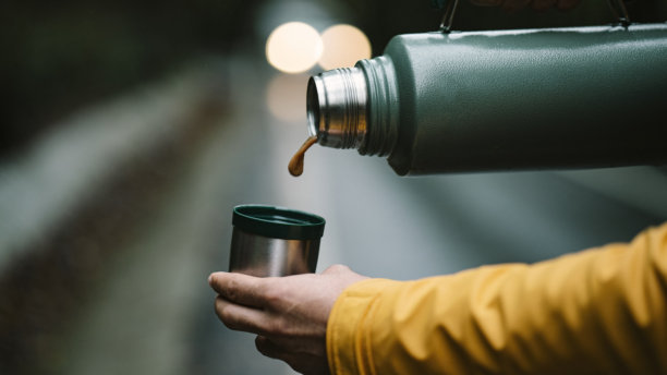 男人的手将咖啡从绿色保温瓶倒入杯子。金属不锈钢保温瓶在森林中的照片。图片下载