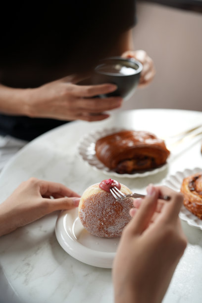 一张裁剪的照片，捕捉到两个朋友在温馨的咖啡馆享用糕点和咖啡的时刻。画面重点在他们的手上，分享美食和交谈。图片下载