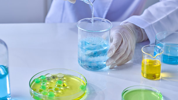 照片从高角度近距离拍摄，白色实验室桌上展示了各种实验室玻璃器皿，里面装有彩色液体和凝胶球，一位科学家正在向玻璃烧杯中倒入蓝色精华。图片下载