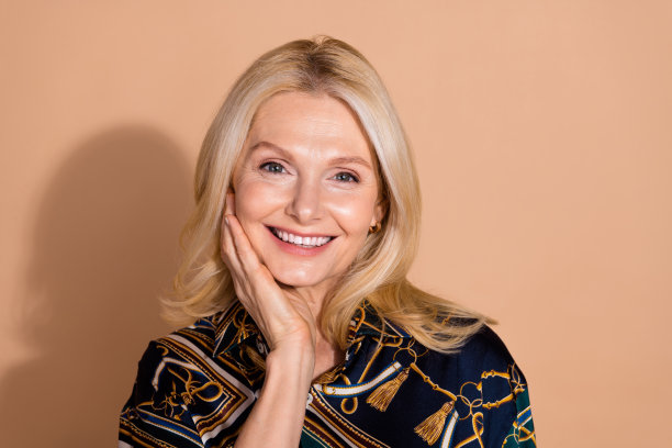 金发快乐的成熟美女模特在米色背景下穿着华丽衬衫，轻触面部肌肤的照片肖像。图片下载
