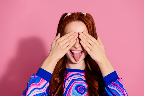 可爱的年轻女士的照片肖像，遮住眼睛，伸出舌头，穿着时尚的蓝色彩色服装，背景为粉色。图片下载