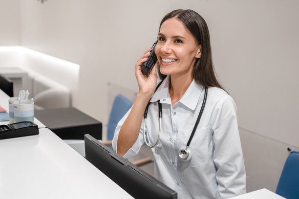 友好的年轻女医生、护士、接待专员穿着白色工作服，坐在办公室里，打电话，与客户交谈，安排预约。图片下载
