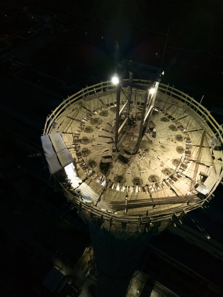 垂直照片。黄昏时分照亮的建筑工地。夜间危险作业。工人在为工厂建造高烟囱。承包商在高层建筑上修理烟囱。工地上的工人戴着安全帽。图片下载