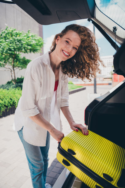 穿着白衬衫的漂亮甜美女性在现代城市户外的汽车后备厢旁拿着行李箱的竖版照片图片下载