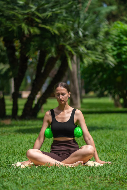 年轻女性坐在公园的地毯上，双腿盘坐，腋下夹着两个带刺的按摩橡胶球。缓解肌肉疼痛，筋膜释放。竖版照片图片下载