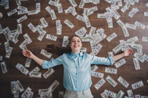俯视照片中，甜美迷人的女经济学家穿着衬衫和眼镜，躺在室内工作场所的地板上数着美元。图片下载