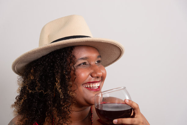 美丽的黑人女性戴着浅棕色帽子，手持玻璃杯饮用葡萄酒。工作室照片。背景为浅色。图片下载