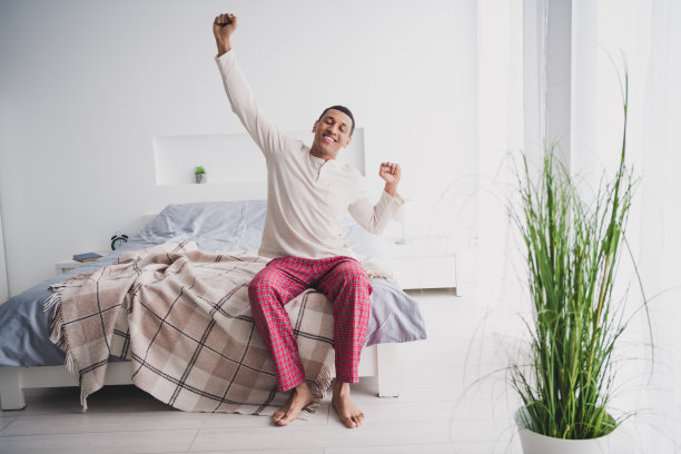 全身照片：一位迷人的年轻男性穿着睡衣坐在床上伸展，享受温馨的阳光，家居室内卧室环境。图片下载
