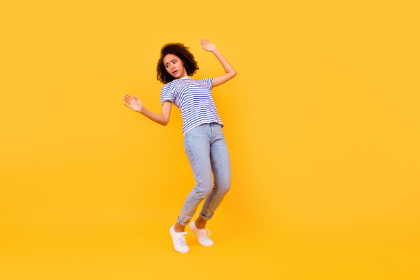 全身照片显示一个紧张的年轻人踮起脚尖，平衡在悬崖边缘的空旷空间，背景为黄色。图片下载