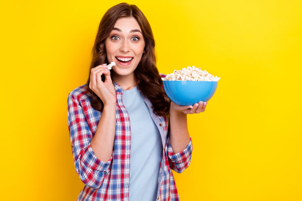 特写照片：年轻女孩兴奋且震惊地吃着爆米花，观看新的家庭剧集，心情愉快，背景为黄色。图片下载