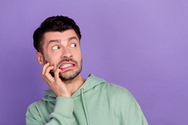 特写照片：年轻、搞笑、兴奋又紧张的男学生穿着卡其色连帽衫，咬着手指，面露恐惧，孤立在紫色背景前。图片下载