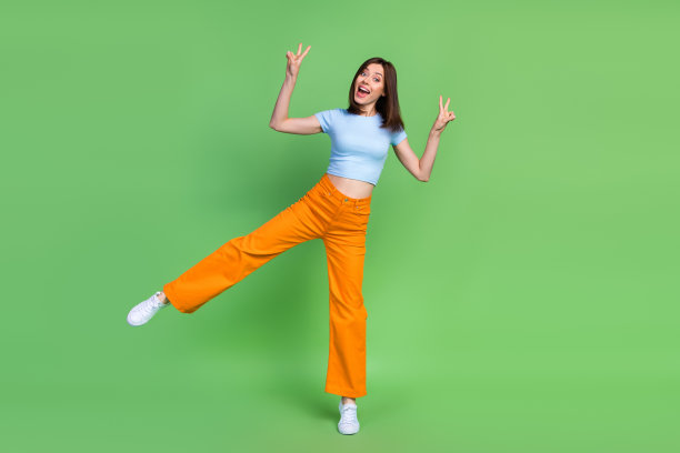 全身照片，快乐的漂亮女孩单腿站立，手指做出V字手势，背景为绿色。图片下载