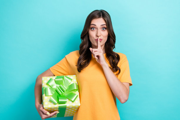 特写照片：年轻可爱女性穿着橙色T恤，做出安静的手势，手持秘密礼物，背景为青色。图片下载