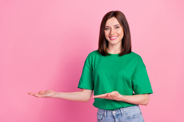 美丽年轻女孩的照片肖像，手掌指向空白处，展示新产品，穿着时尚的绿色服装，背景为粉色。图片下载