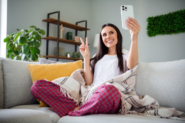 漂亮年轻女士在现代公寓室内穿着睡衣，拍摄自拍并做出V字手势的全身照片。图片下载