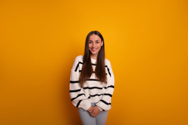 这张图片展示了一位微笑的年轻女性自信地站在明亮的橙色背景前。她穿着白色条纹毛衣和浅蓝色牛仔裤，双手合十，姿势显得很随意。图片下载