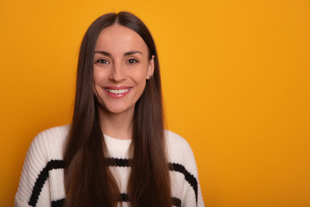 这张图片展示了一位微笑的年轻女性特写，她有着长长的棕色头发，站在明亮的橙色背景前。她穿着一件白色条纹毛衣，散发出温暖和积极的气息。图片下载