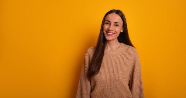 一位长着棕色长发的快乐女性在鲜艳的黄色背景下灿烂微笑。她穿着米色毛衣，散发出温暖、积极和友好的气息，这是一张摄影棚拍摄的照片。图片下载