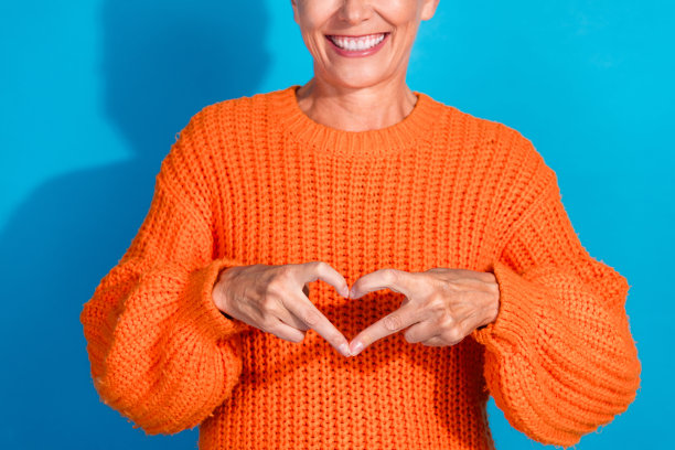 穿着橙色毛衣的老太太在蓝色背景前做出心形爱心符号的裁剪照片图片下载
