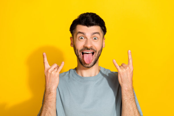 特写照片：年轻有趣、无忧无虑、心情愉快的男人，做出摇滚爱好者的手势，舌头伸出，背景为黄色。图片下载
