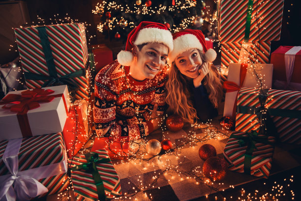 年轻情侣穿着圣诞服装，躺在地板上享受温馨的圣诞派对装饰，周围是购物礼物的全身照片。图片下载
