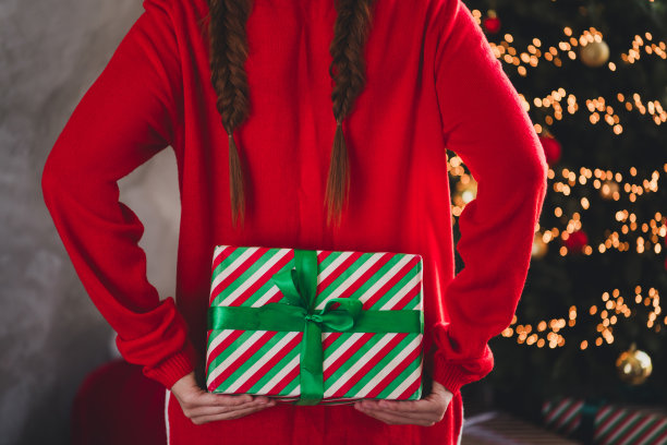 年轻的红发女性在家中派对装饰的室内，穿着圣诞服装，背后藏着礼物，惊喜的剪裁照片。图片下载