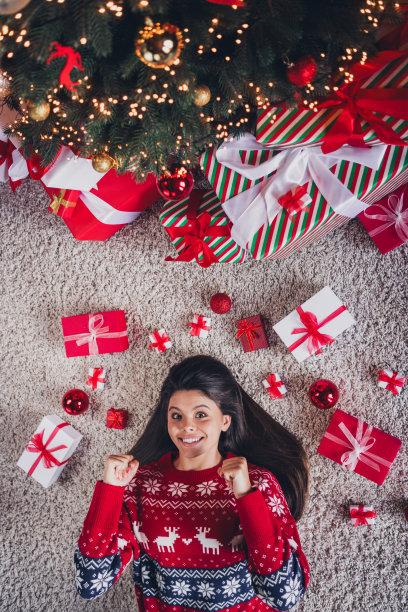 高角度拍摄可爱女孩穿着新红色印花衣服，躺在地毯上享受室内圣诞节装饰的照片。图片下载