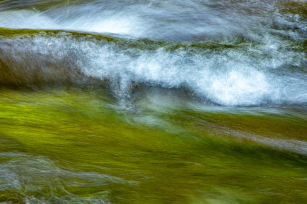 一张长时间曝光的照片，展示了一条河流在岩石上流动，水面模糊成平滑的图案。周围树木的绿色叶子在水中倒映。图片下载