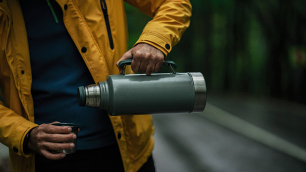 穿着黄色夹克的男子正在将饮料从保温瓶倒入玻璃杯。照片中男子在森林里拿着金属不锈钢保温瓶。图片下载