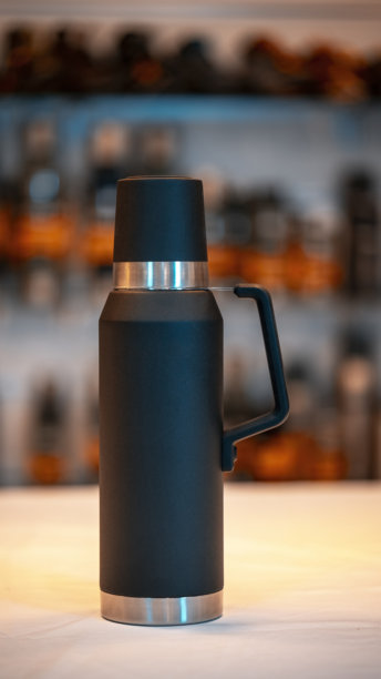 黑色真空不锈钢保温瓶放在白色桌子上的单张照片。侧面带把手的保温瓶特写照片。图片下载