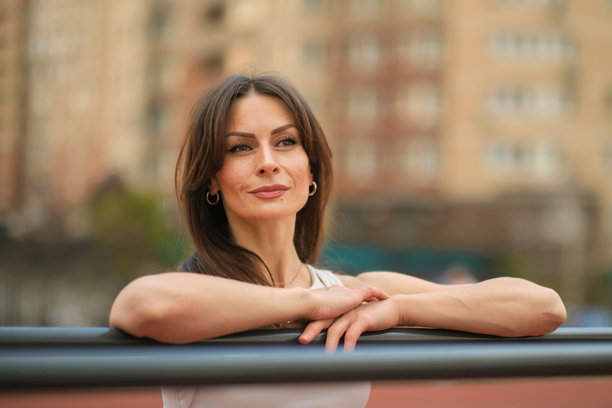 一位自信的女性倚靠在健身器械上，面带温暖的微笑，背景是城市风景。这张图片突显了积极的心态和从积极生活方式中获得的自信。图片下载