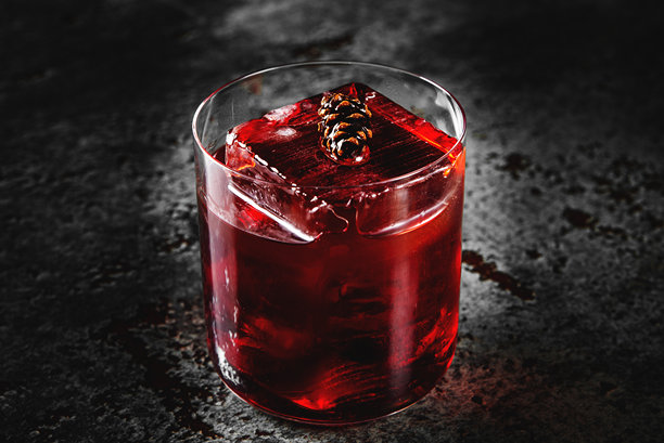 一杯充满活力的红色鸡尾酒，杯中放有冰块，装饰着松果，置于深色纹理表面上。非常适合节日和冬季主题的库存照片。图片下载