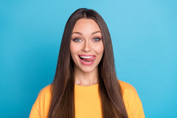 特写照片，漂亮积极的女士满意地舔着牙齿，佩戴着牙套，穿着黄色衣服，背景为蓝色。图片下载