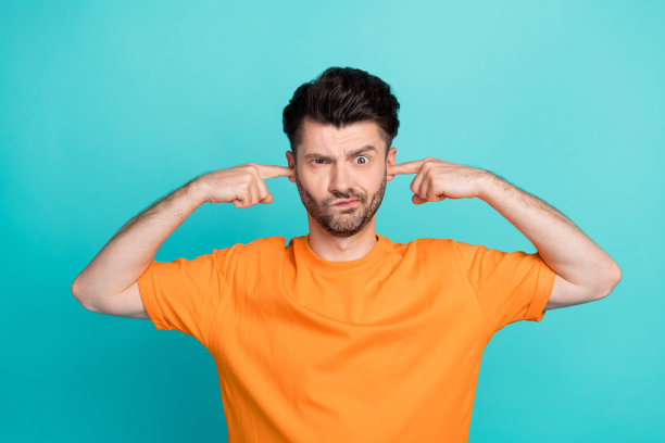 特写照片：年轻有趣、迷人、帅气的可爱男孩穿着橙色T恤，双手捂住耳朵，撅嘴不满，因大声音乐而做出鬼脸，背景为青色。图片下载