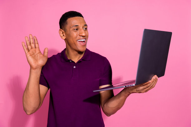 年轻男性的照片肖像，手持笔记本电脑进行视频通话，挥手致意，穿着时尚的紫色服装，背景为粉色。图片下载