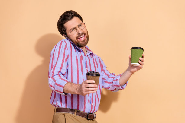 年轻男性的照片肖像，因电话交谈而感到烦恼和不快，手持咖啡，穿着时尚的条纹正装，背景为米色。图片下载