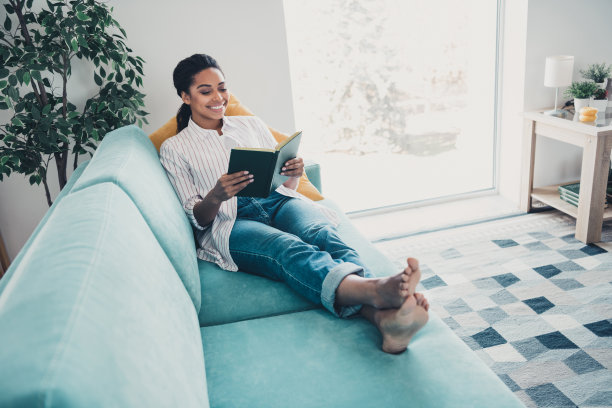 漂亮年轻女孩在现代公寓室内沙发上穿着衬衫阅读书籍的全身照片图片下载