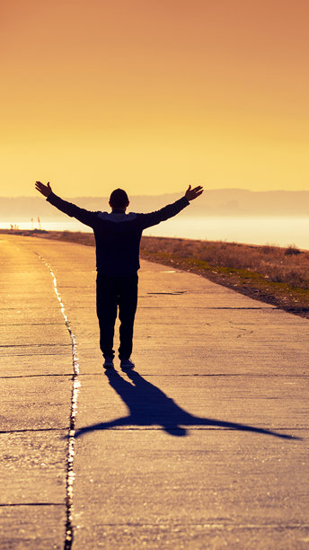 一位年轻男子双手举起，走在混凝土道路上，欣赏美丽的橙色日落。竖版图片图片下载