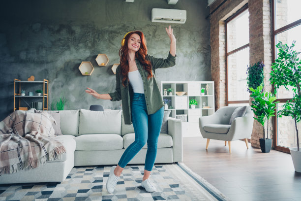 可爱的年轻红发女孩在宽敞的室内房间里穿着卡其色衣服，戴着耳机跳舞，心情愉快的全身照片。图片下载