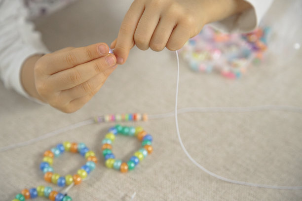 孩子的手特写，正在将五颜六色的珠子穿到绳子上，专注于制作手工手链。这张图片展现了创造力、精细运动技能和有趣的学习。图片下载
