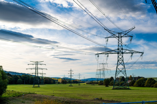 一幅风景照片，展示了电力线和输电塔延伸穿过开阔的田野，背景是丘陵和部分多云的天空。该图像捕捉了自然与工业的交汇，突显了基础设施在乡村中的作用。图片下载