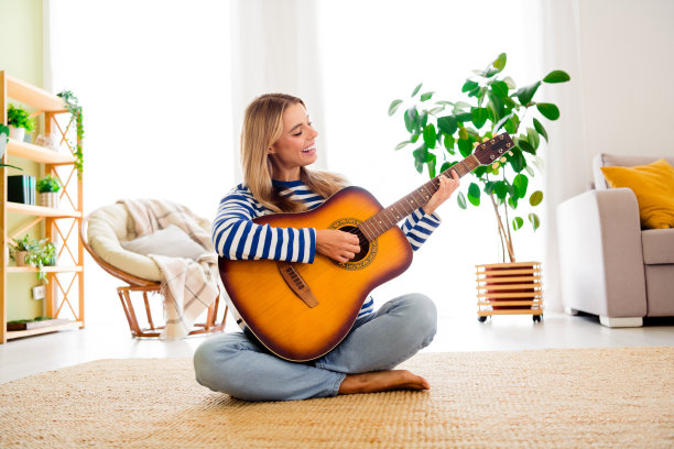 全身照片，漂亮的金发年轻女孩坐在地板上弹吉他，穿着条纹衣服，享受现代舒适的室内公寓环境。图片下载