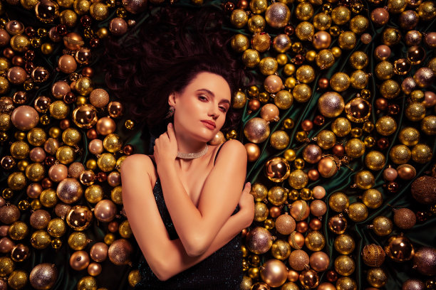 俯视照片中，时尚女士抱着自己，感到诱惑，躺在金色圣诞装饰上，穿着华丽的裙子。图片下载