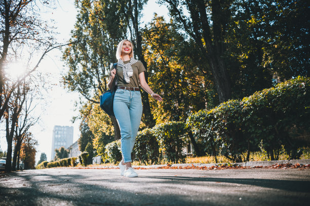 年轻漂亮的金发女子在城市公园外穿着卡其色衣服，肩上搭着毛衣，走路的全身照片图片下载