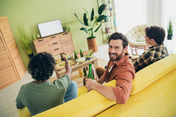 公司三位朋友在现代室内公寓里聚会，喝啤酒，看电视，享受周末时光的背面照片图片下载